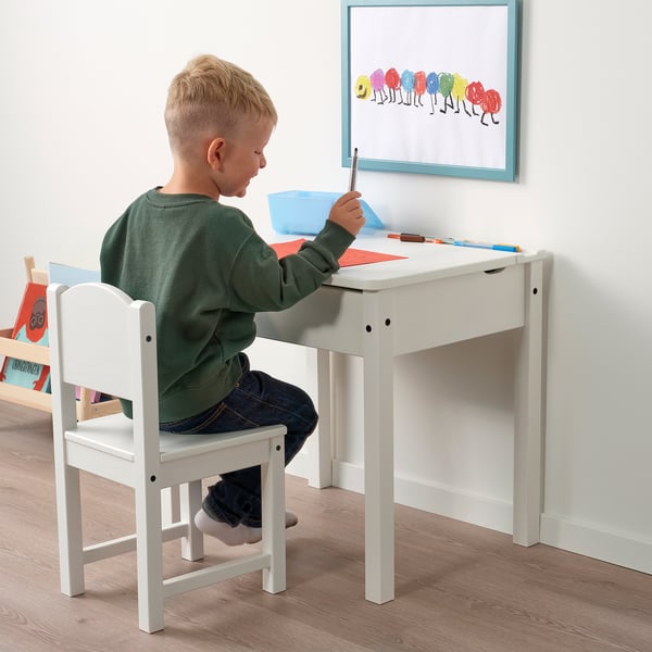 Child sitting at small white desk, drawing. Desk has lift-up lid and storage. Picture on wall above.