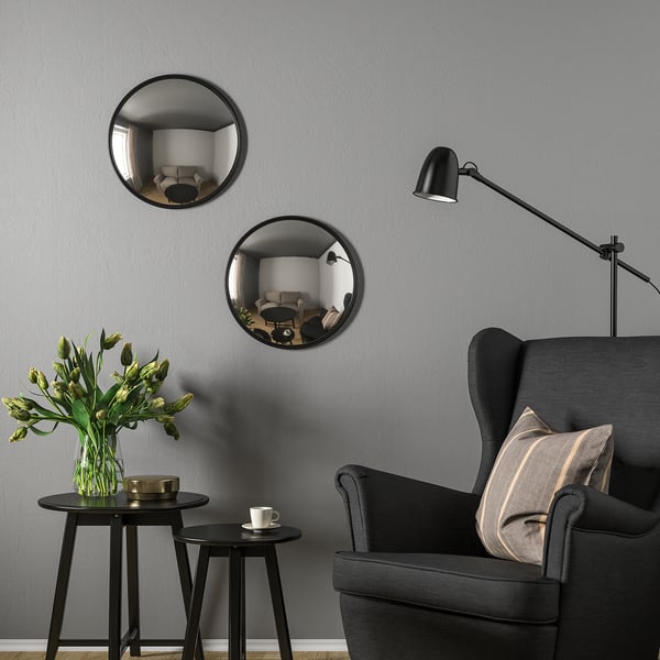 Black mirrors SVARTBJÖRK on grey wall, reflecting living room. Paired with black chair and small tables - one with flowers, one with cup.