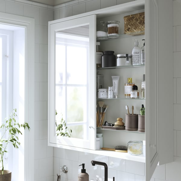 A white bathroom cabinet with mirrored doors, featuring interior shelves stocked with toiletries and beauty items, mounted above a sink.