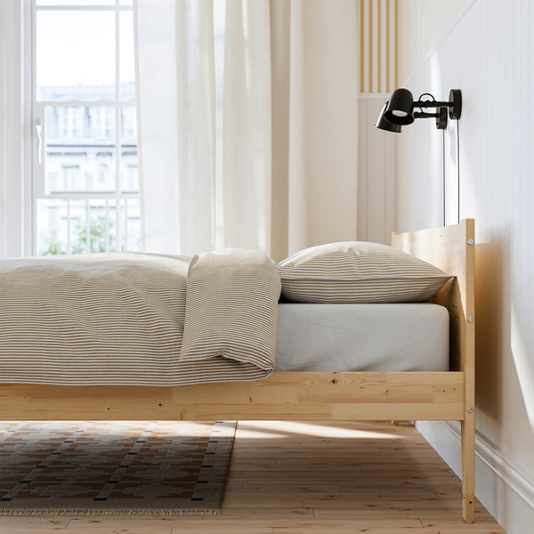 Minimalist wooden bed frame with striped bedding, mounted black reading lamp, and slatted bed base for air circulation.