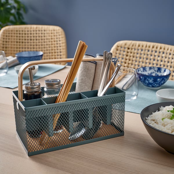 A VIMPELFISK green metal cutlery caddy with bamboo handle, holding cutlery and napkins, placed on a wooden table with a bowl of rice.