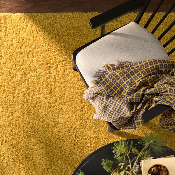 Bright yellow ÄRENDE rug, softer than chair, covers floor widely, with dark chequered blanket and small plant nearby.