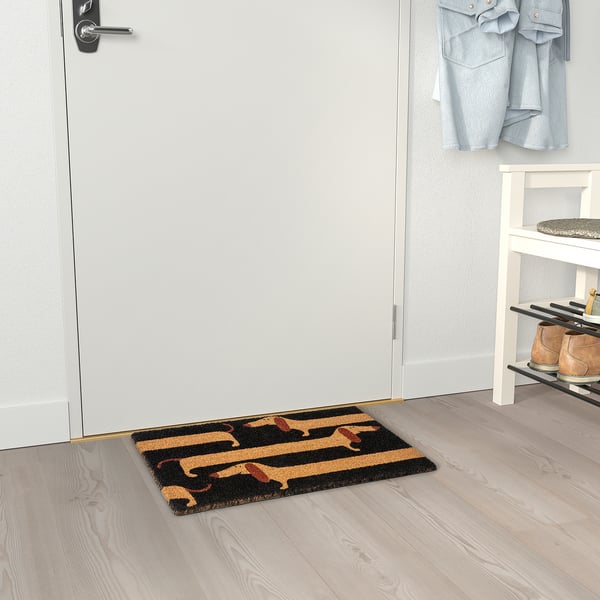 Door mat with black and tan dachshund design lies near a white door and shoe rack.