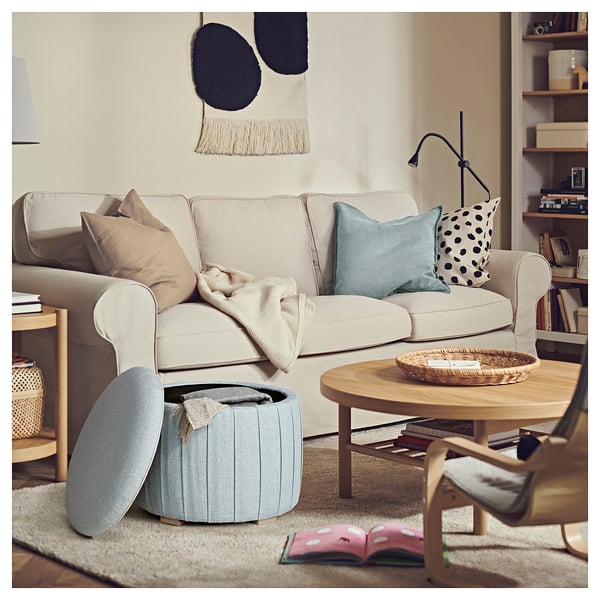 Living room: beige sofa with pillows, blue pouffe, wooden table, books, and shelves.