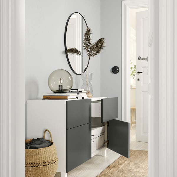 Modern entryway with BESTÅ storage in white and dark grey. Open shelves, drawers, and a round mirror. Woven basket and decorative items are included.