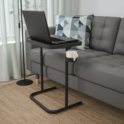 Black laptop stand with mug on steel base next to gray sofa.