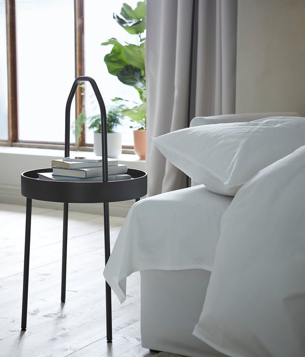 Bedside table BURVIK, black, round tray top, curved handle, white bed linens, indoor plant by window.