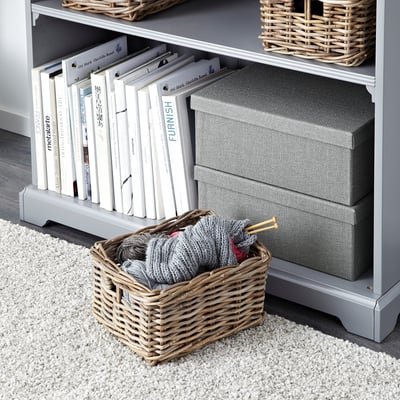 A wicker basket on a rug, filled with yarn and knitting needles, beside a grey bookcase with books, woven baskets, and storage boxes on shelves.