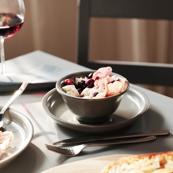A bowl of ice cream topped with whipped cream and berries sits on a plate. Beside it, a wine glass and a fork are neatly arranged on a table.