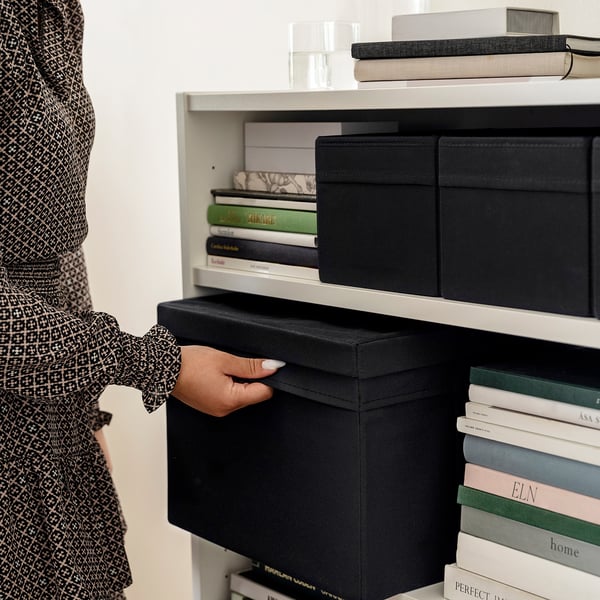 A person in patterned clothing pulls a black fabric lid off a box on a white shelf, which holds more boxes and books.