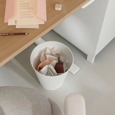 Small white FNISS bin under desk, holding crumpled papers, next to a beige chair and part of a wooden desk with a pen and sticky notes.