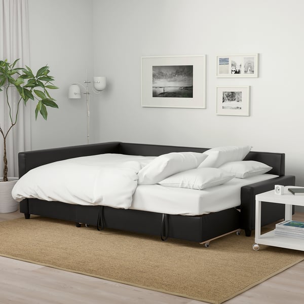 Black FRIHETEN sofa-bed with white pillows in minimalist room. Photos, plant, side table with magazines. Light rug, wooden floor, grey curtains.