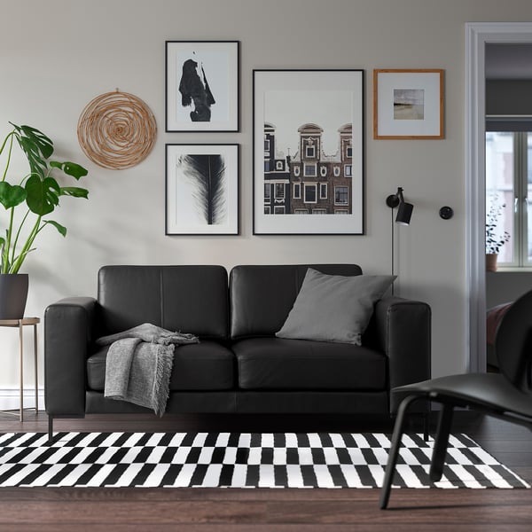 Modern living room with black sofa, grey pillows, art, chequered rug, lamp, plant.