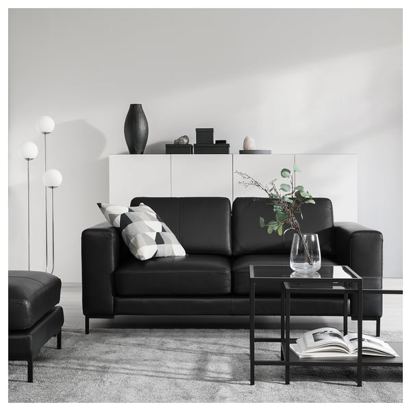 Modern living room with black leather FULLERÖ sofa, geometric throw pillows, glass coffee table with plant and magazines.