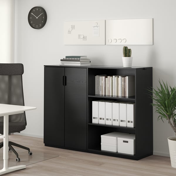 A GALANT black office storage unit with closed and open shelves holding books, binders, and a plant. Next to it, a white chair is visible.