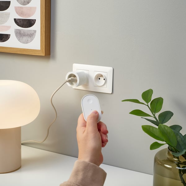 A hand uses a remote to control a lamp plugged into a smart socket. Beside a houseplant and wall art.