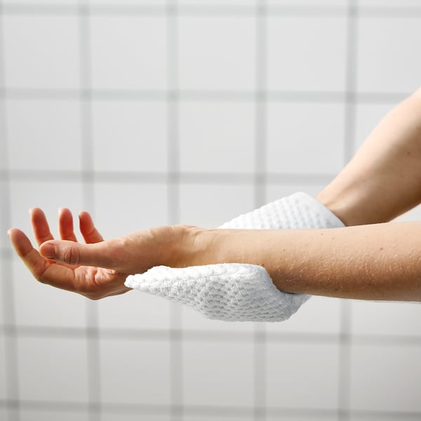 Person washing arm with white sponge, against white tiled wall. Simple hygiene routine.