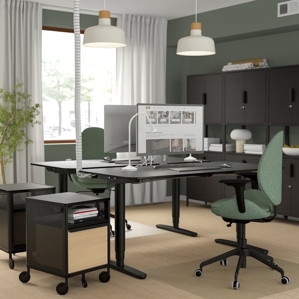 Modern office featuring HATTEFJÄLL green ergonomic chair, black adjustable desk, and hanging lamps. Dark cabinets and large windows.