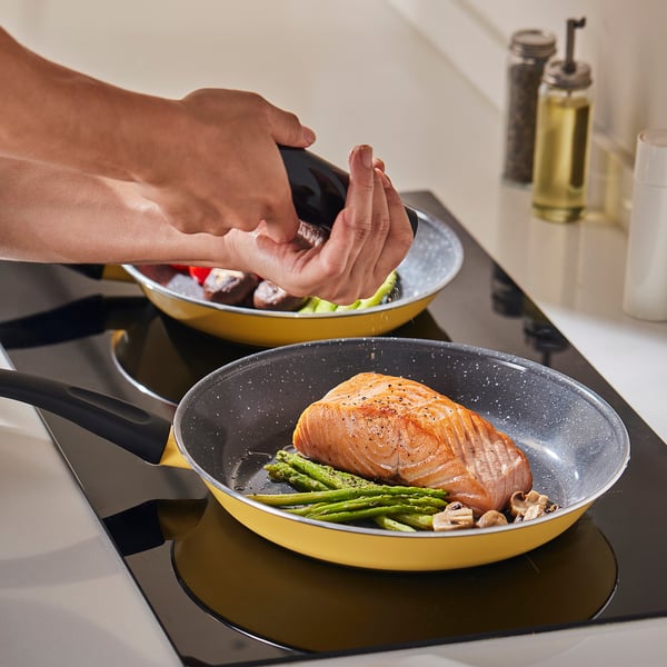 Person seasoning salmon in a yellow non-stick frying pan with green asparagus. Two pans cooking on a stove.