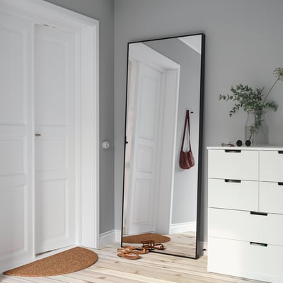 Hövet mirror leans against wall, reflecting corridor. Black frame complements modern decor, surrounded by wooden floor and red handbag.