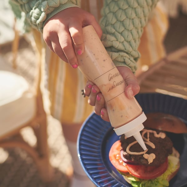 A person squeezes yellow sauce on a burger with a blue plate. They wear a green sweater and pink nail polish.