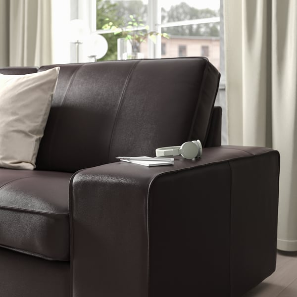 A dark KIVIK sofa with pillows, headphones, and magazines on the armrest by a large window, showcasing its comfortable and functional design.
