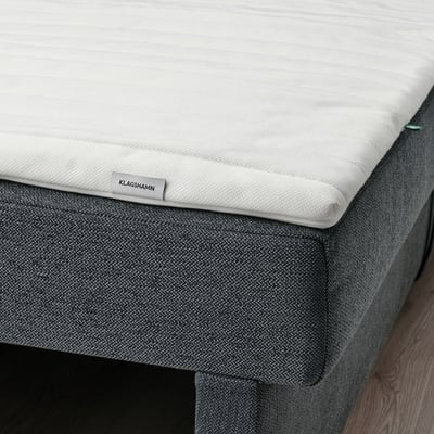 A close-up of a rolled mattress pad labelled KLAGSHAMN on a dark bed base, showcasing its foam filling and stretch fabric designed for comfort.