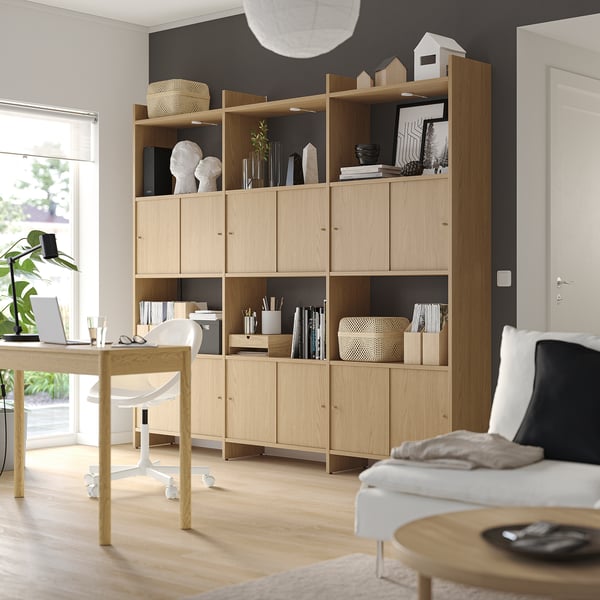 Light wood storage unit with open/closed shelves, holding decor, books, office supplies against dark wall.