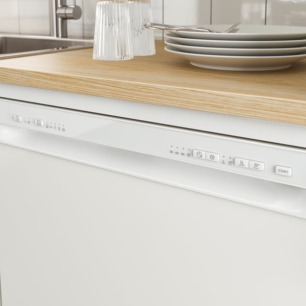 Modern kitchen with white built-in dishwasher under wooden countertop. Control panel visible, cutlery and plates stacked on counter.