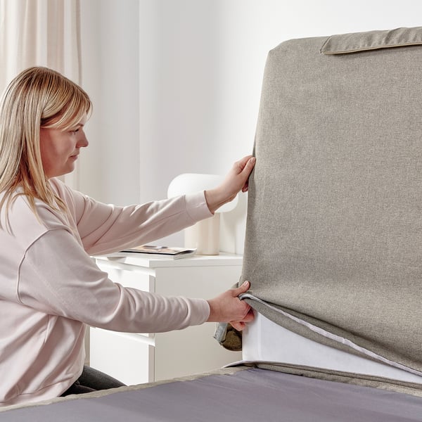 A person in a pink shirt adjusts a grey bed cover with a herringbone pattern, showcasing the easy-to-clean cover for a LILLFJÄLLET divan bed.