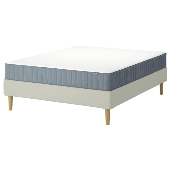 An IKEA LYNGÖR bed base with a white valevåg mattress and beige upholstery. The bed has light wooden legs and a simple, modern design.