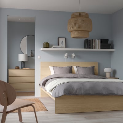 Blue bedroom with oak MALM bed, grey linens, dresser, shelves, 2 lamps, chair. Bed is focal point.