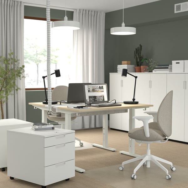 White height-adjustable desk with light wood top, metal base, lamp, filing cabinet, and grey chair.
