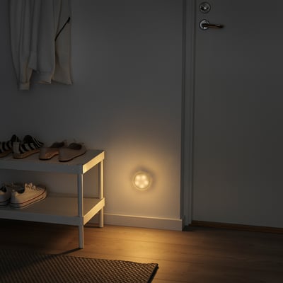 Dark hallway with wall-mounted MOLGAN light, shoe rack and hanging jackets. Light is small, round, and white, illuminating floor.