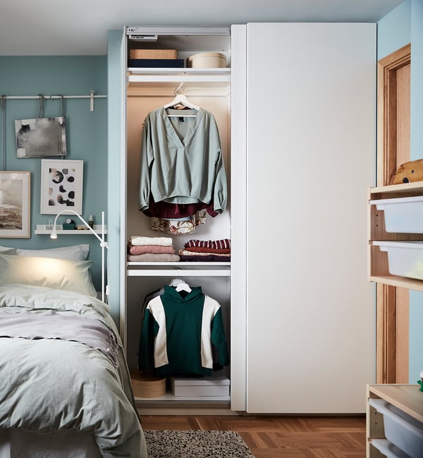 A modern bedroom a PAX wardrobe in white, holding blue and green clothes, with a bed and artwork.