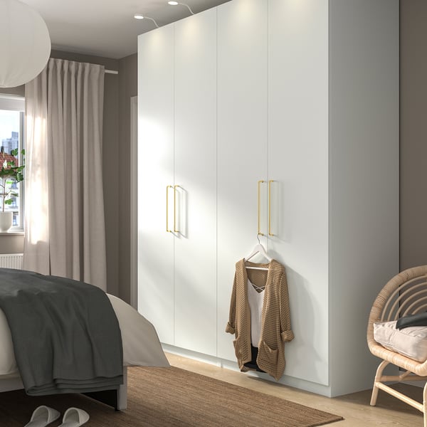 Modern bedroom with white PAX wardrobe, gold handles, sweater, dark/light linens bed, wicker chair near sheer curtains.