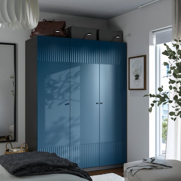Modern bedroom with tall blue wardrobe, high ceiling, & natural light. Wardrobe has sleek handles & storage boxes.