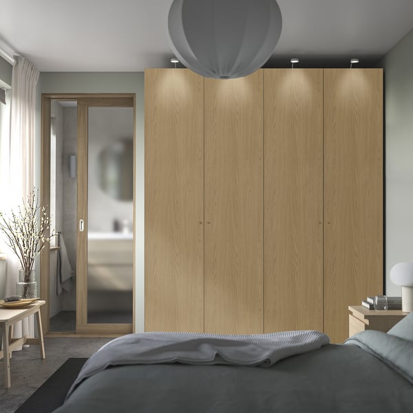 Modern bedroom with large light-wood PAX wardrobe, soft grey bedding, round pendant lamp, and white vessel lamp on wooden bedside table.