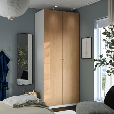 Cosy bedroom with light wooden PAX wardrobe, blue wall, grey mirror, and vibrant art. Comfortable with white rug and grey armchair.