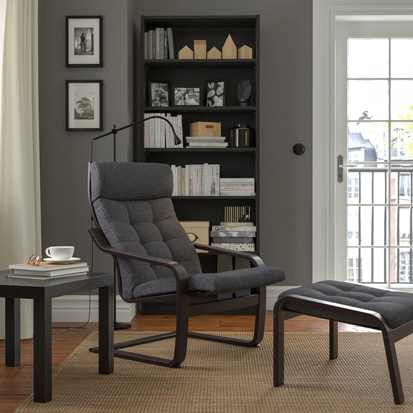 Modern living room with black POÄNG armchair, footstool, side table, bookshelf, and window. Dark grey upholstery, wooden frame, cosy setup.