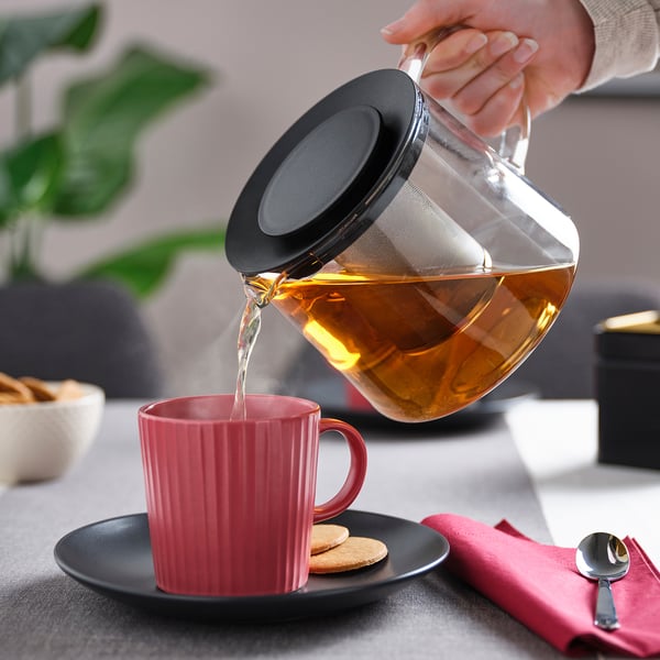 Teapot pours tea into red mug on dark plate, with integrated infuser, steam rising from spout.