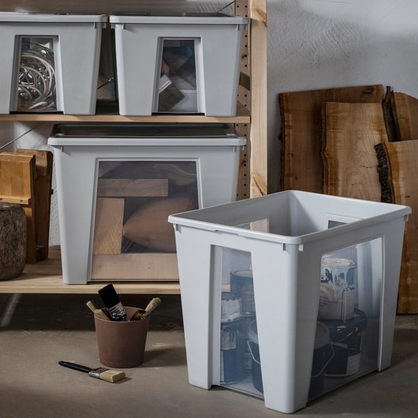 White SAMLA storage box with clear windows holds paint cans, placed on floor near wooden shelves holding more SAMLA boxes, wood, and tools.