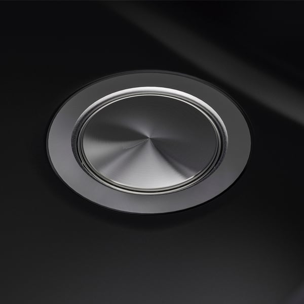 Round stainless steel sink drain cover on black sink. High-quality, reflective surface with smooth finish. Centred, polished metallic design.