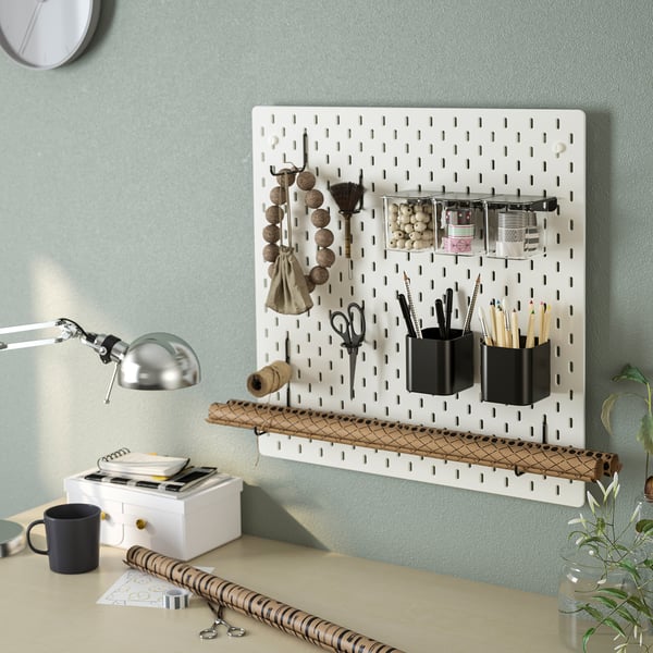 SKÅDIS pegboard organiser on green wall holds cups, brushes, scissors, containers, and a lamp above desk.