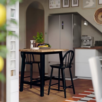 Small black SKOGSTA bar table with chairs, wooden top, in kitchen with refrigerator, countertop appliances, and wall decor.