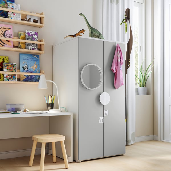 Kids room with light grey SMÅSTAD wardrobe, white desk, wood stool, lamp, books, toys, and plants.