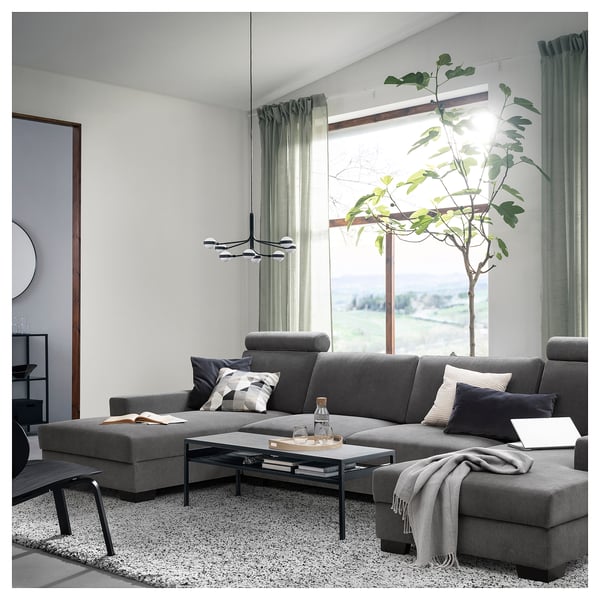 Modern living room with large grey l-shaped SÖRVALLEN sofa. Decorated with pillows, rug, and a tall plant near large windows.