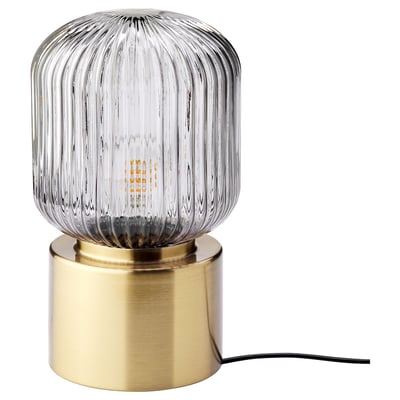 A SOLKLINT lamp with a brass base and grey glass shade. Its cylindrical shape is accented by vertical glass lines, emitting soft, warm light.