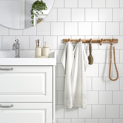 Modern bathroom with STOREDAMM wooden hook rack holding white robe. White tiles and cream dispensers. Acacia wood.