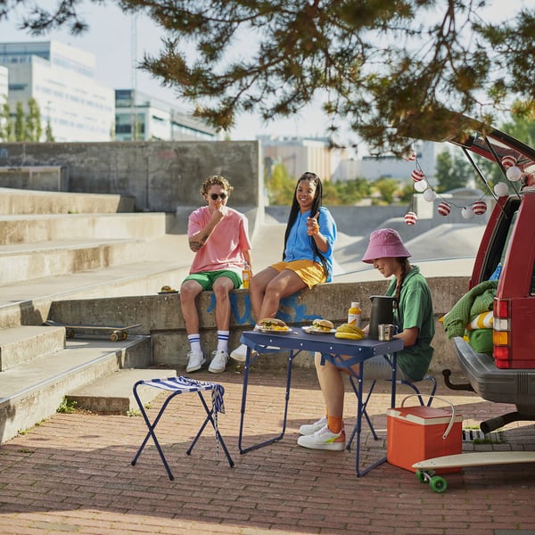 Three people sit by colourful STRANDÖN outdoor furniture at an urban picnic.